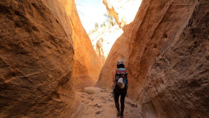 Hiker with helmet and gear walking through narrow sandstone slot canyon in Birch Hollow, Zion area