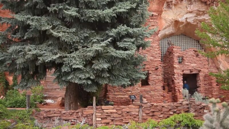 Small red stone structure built into Moqui Caverns with pine tree in front