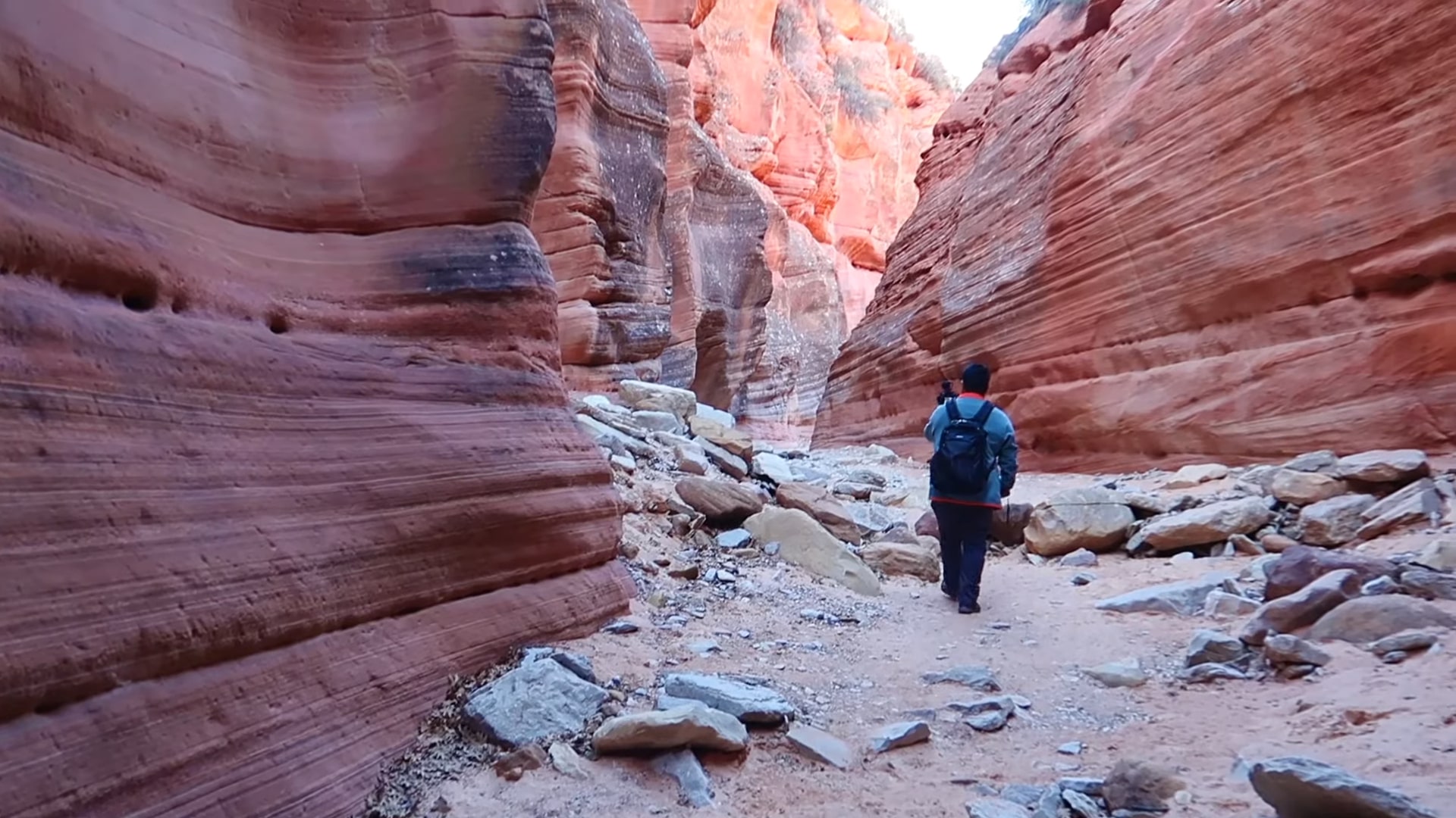 A hiker with a backpack walking through the narrow, red rock walls of Peekaboo Slot Canyon, surrounded by rugged rock formations and scattered stones