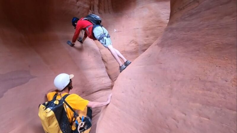 Two canyoneers climbing slick red rock walls inside Upper Red Cave in Utah