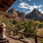Two people in hats sitting on a wooden porch, overlooking Zion National Park's red rock cliffs and forested valley