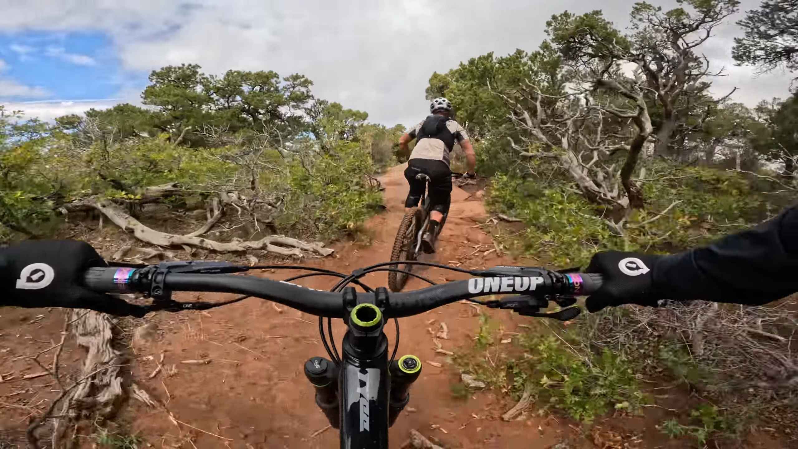 First-person view of two mountain bikers riding a rugged desert trail surrounded by brush and trees