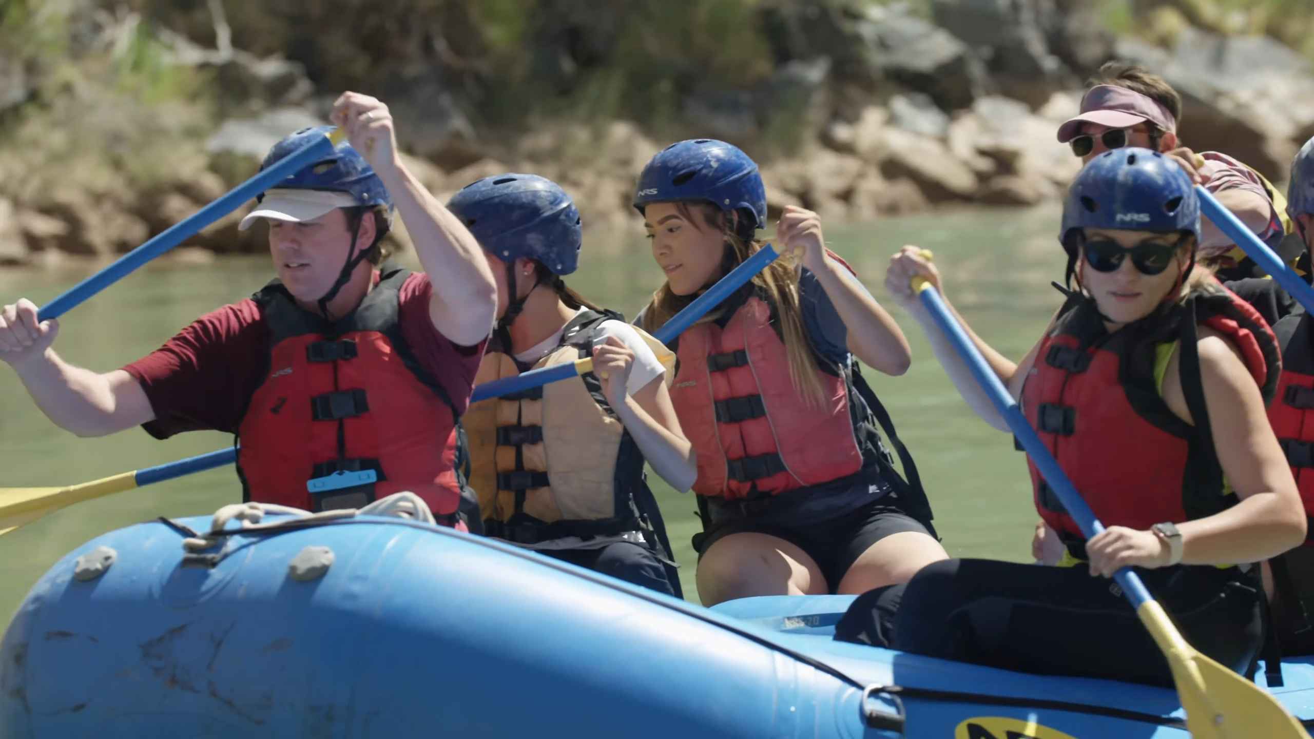 A group of people wearing helmets and life jackets paddling together in a blue raft on a river