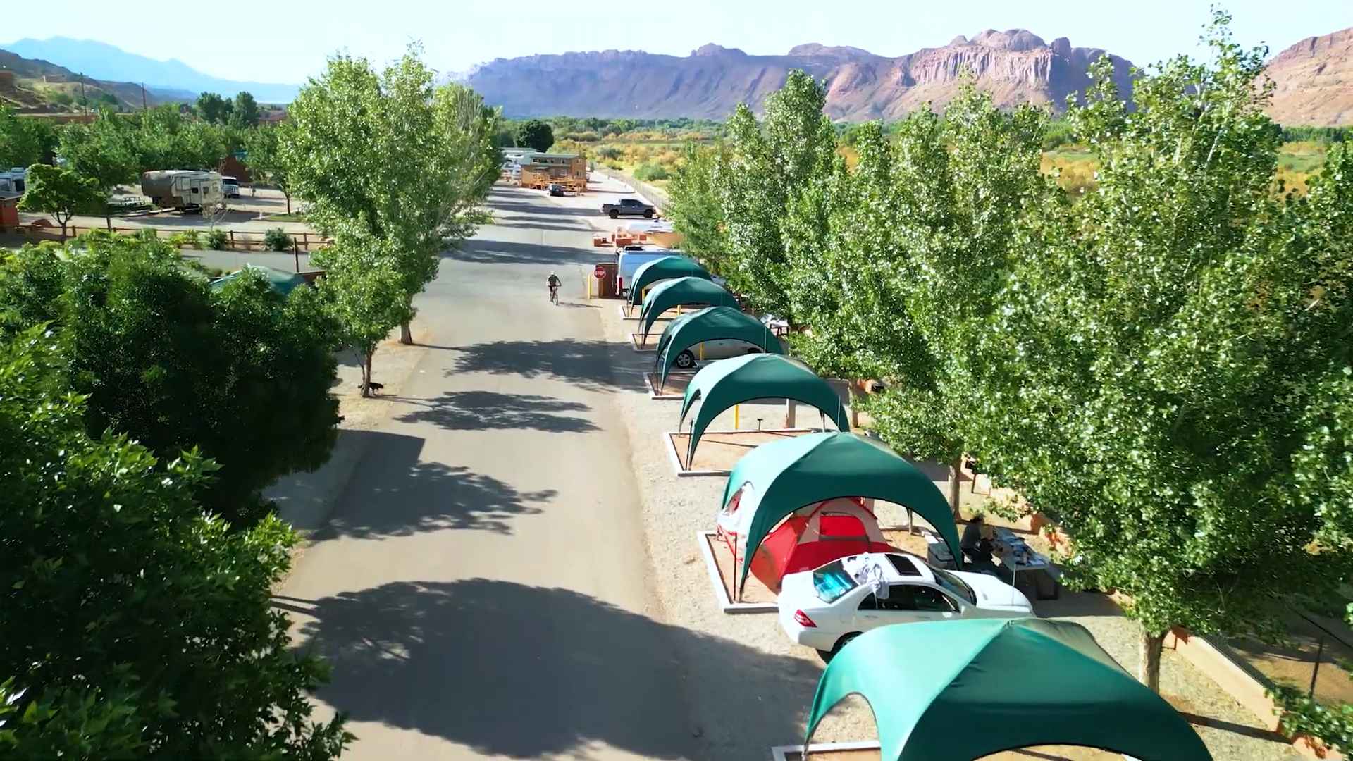 A row of shaded tent sites and parked cars at a campground with mountain views in the background