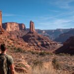 A person with a backpack stands on a desert trail overlooking towering red rock formations and canyons in Moab, Utah