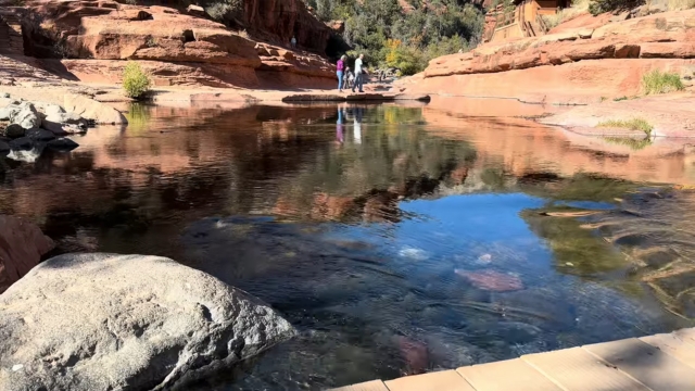 A calm natural pool surrounded by Sedona’s red rock formations with people walking nearby