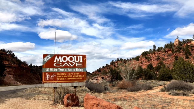 Sign for Moqui Cave Museum along the road with red rock cliffs and trees in the background