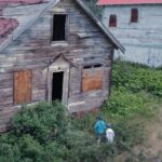 Two children explore a weathered wooden house, overgrown with vegetation