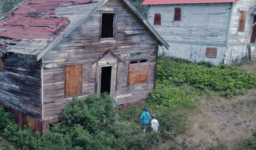 Two children explore a weathered wooden house, overgrown with vegetation