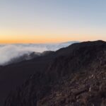 Rocky volcanic slopes stretch above the clouds at sunset, offering a dramatic view from a high-altitude hiking trail