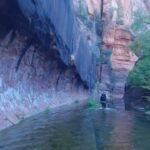 A hiker walks through shallow water beneath towering red rock cliffs and trees on one of the best Sedona hikes