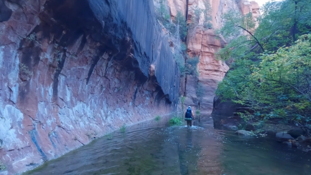 A hiker walks through shallow water beneath towering red rock cliffs and trees on one of the best Sedona hikes