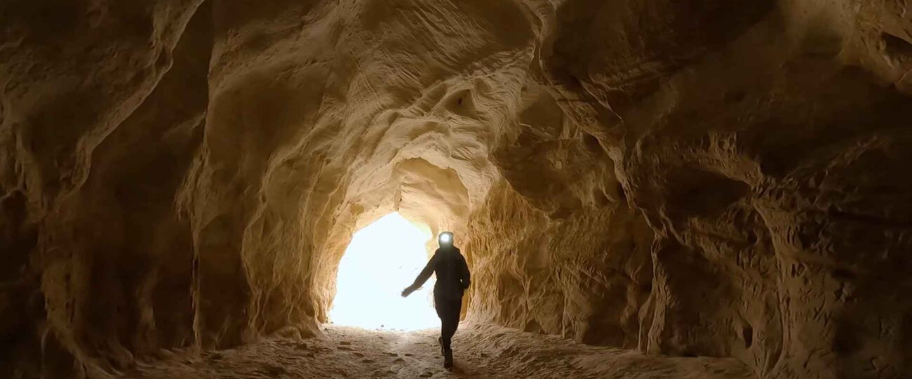 A hiker walking through the Belly of the Dragon tunnel in Utah, silhouetted against the bright light at the tunnel’s exit, surrounded by textured sandstone walls