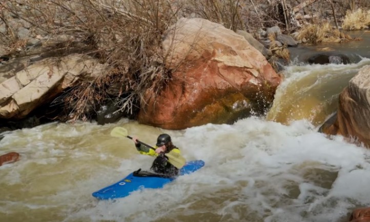 A man kayaks through a river with visible rocks and rapids
