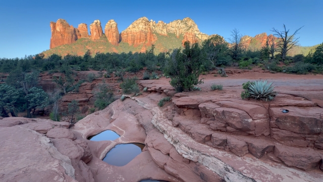 Small pools of water rest on red sandstone with Cathedral Rock glowing in the background at sunset