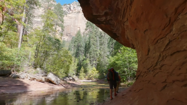 A hiker walks along a shaded red rock wall beside a shallow creek with tall cliffs and pines in the background