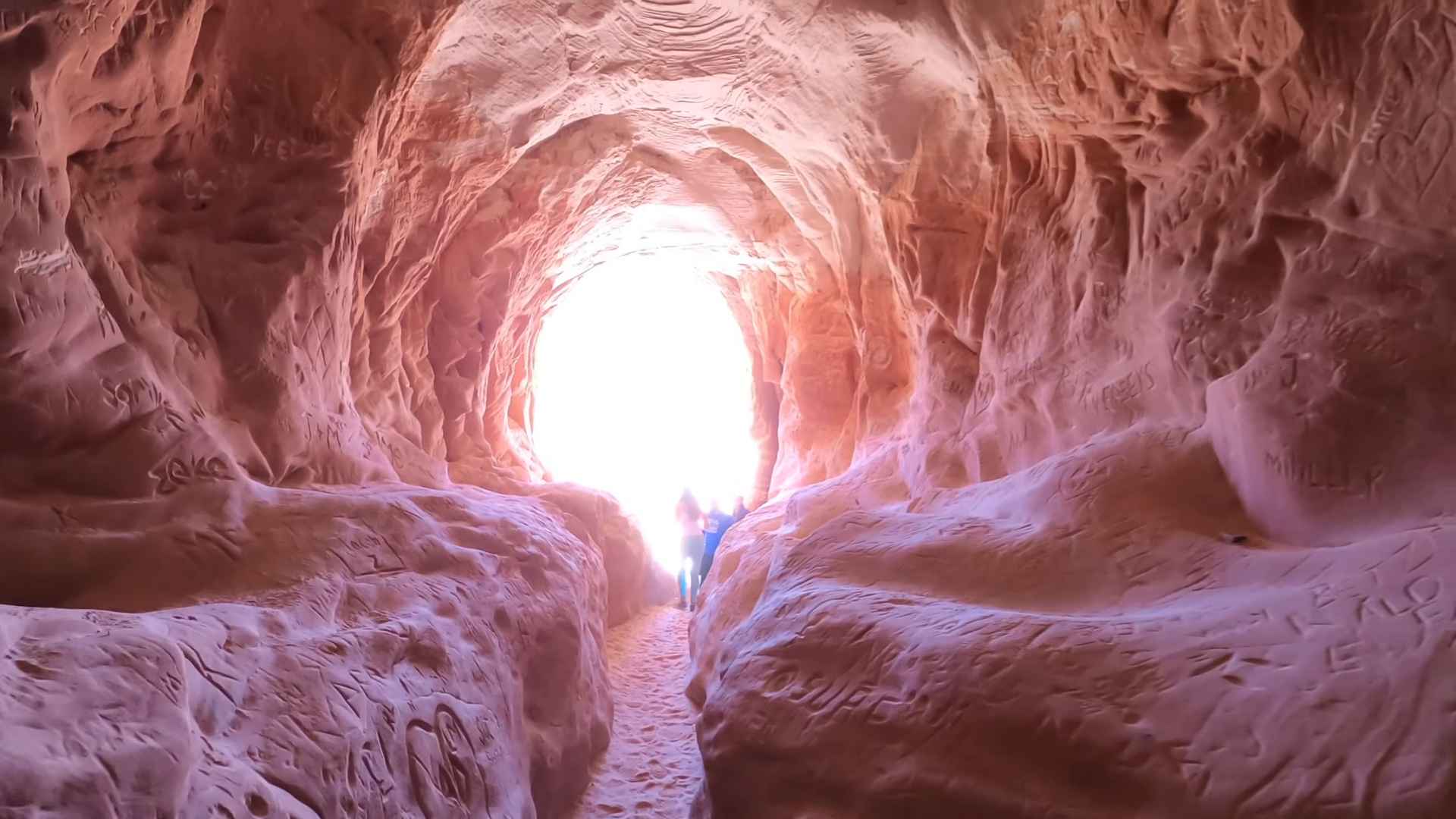 Hikers walking through the Belly of the Dragon tunnel in Utah, a red sandstone cave-like passage glowing with sunlight at the entrance