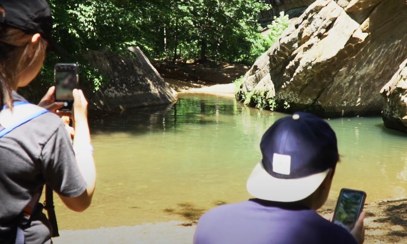 Two people photographing a river with their phones, capturing the scenic view and natural surroundings