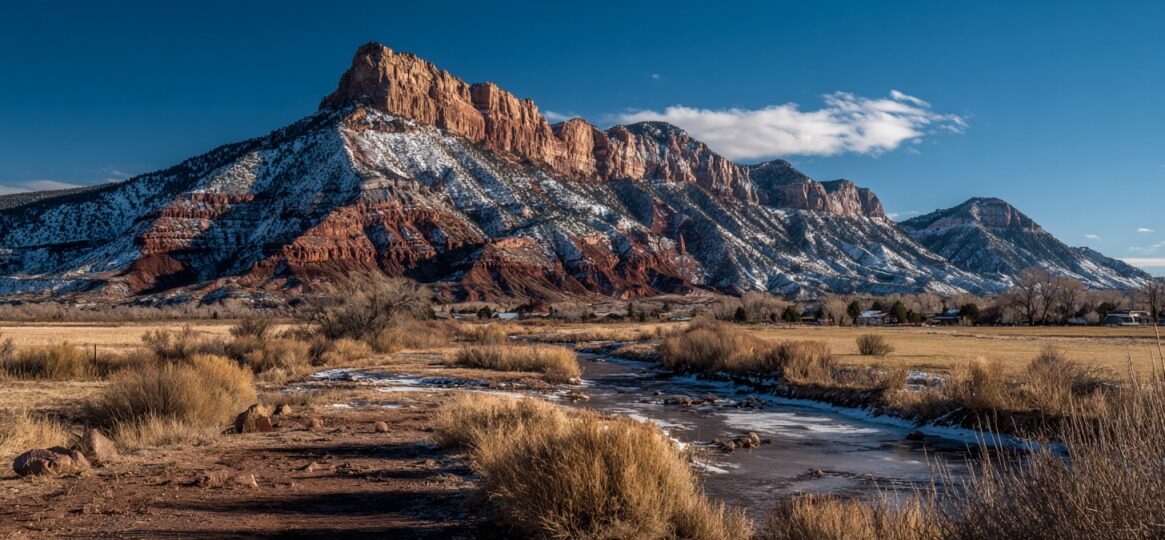 Scenic landscape of Mt. Carmel, Utah with red cliffs, snow-dusted mountains, dry grass, and a winding stream under a clear blue sky