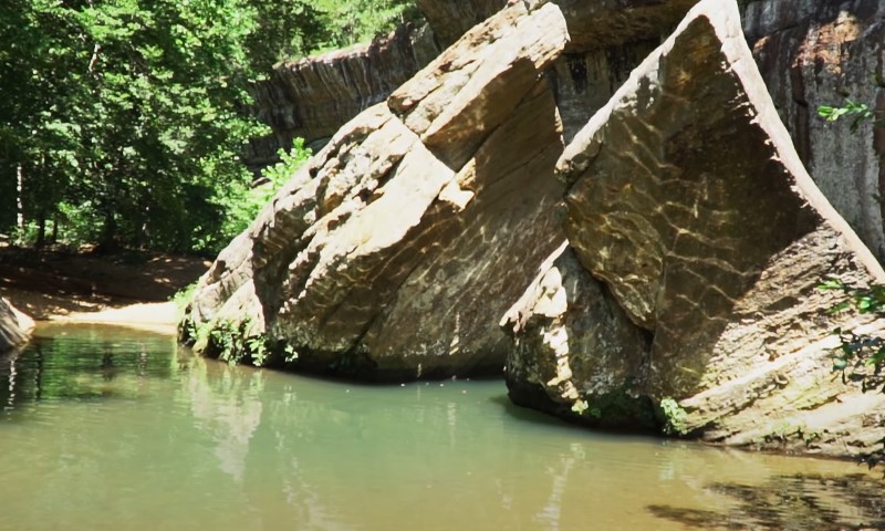 Two massive, sunlit boulders rise sharply from the emerald green water of a shaded forest pool.