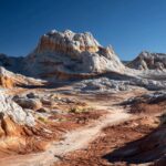 Shallow water reflecting layered red, orange, and white rock formations under a bright blue sky at White Pocket, Arizona
