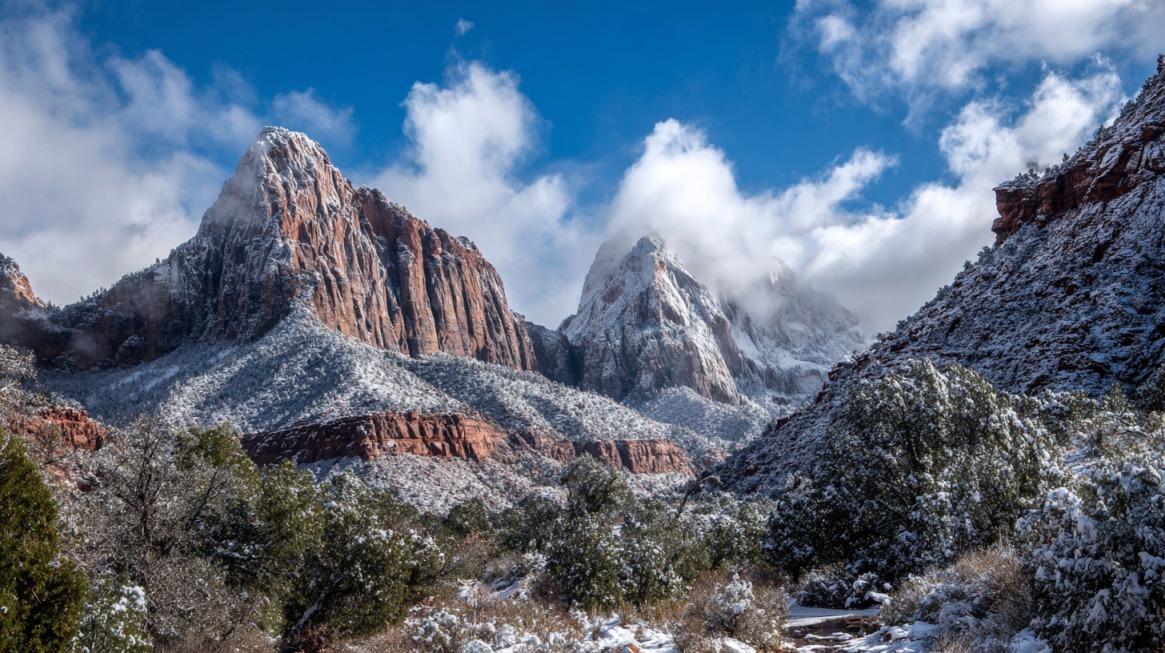 Snow covered red rock mountains and trees under a partly cloudy blue sky
