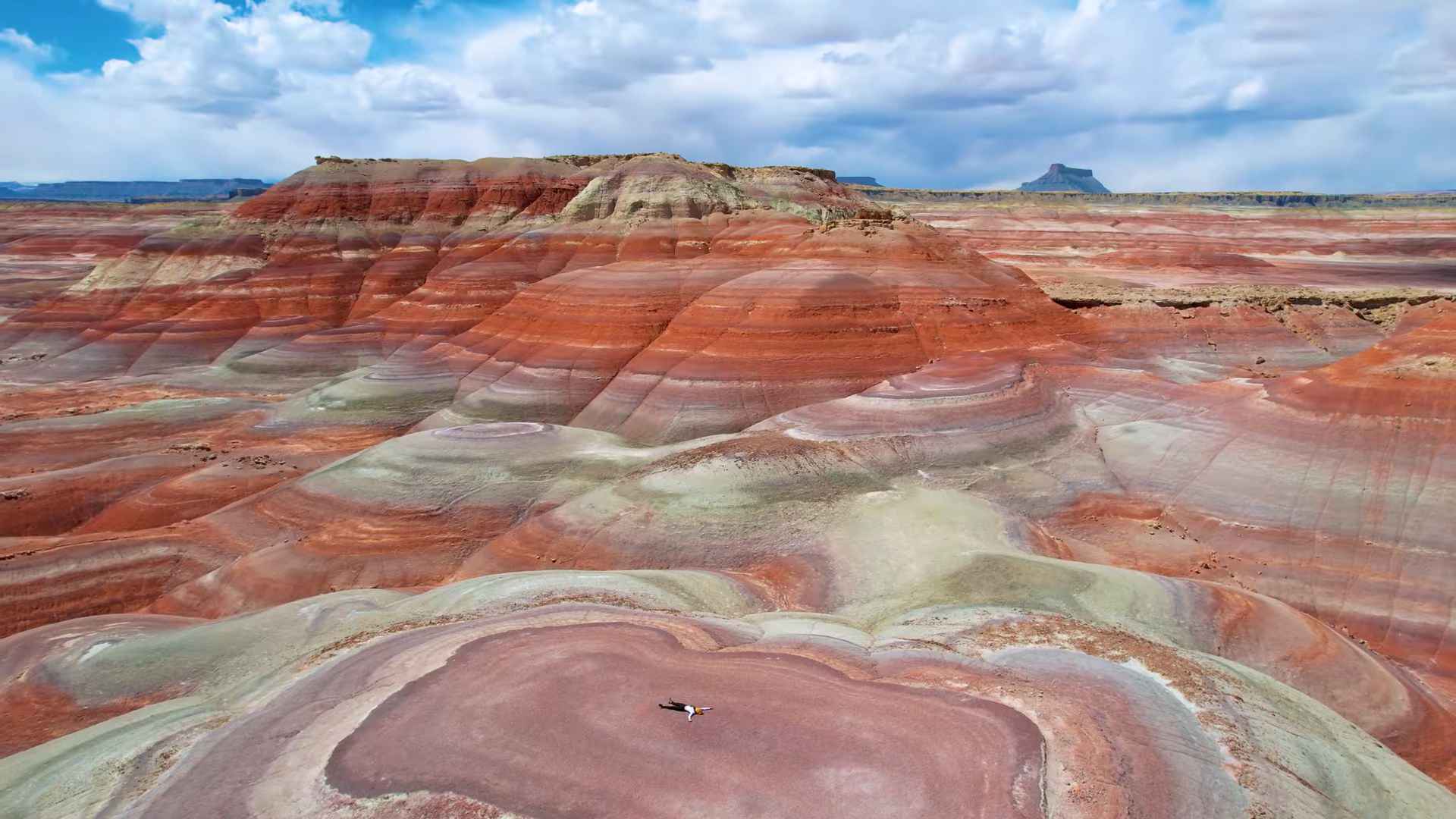 Person lying on layered red, pink, and gray clay hills in a wide desert landscape