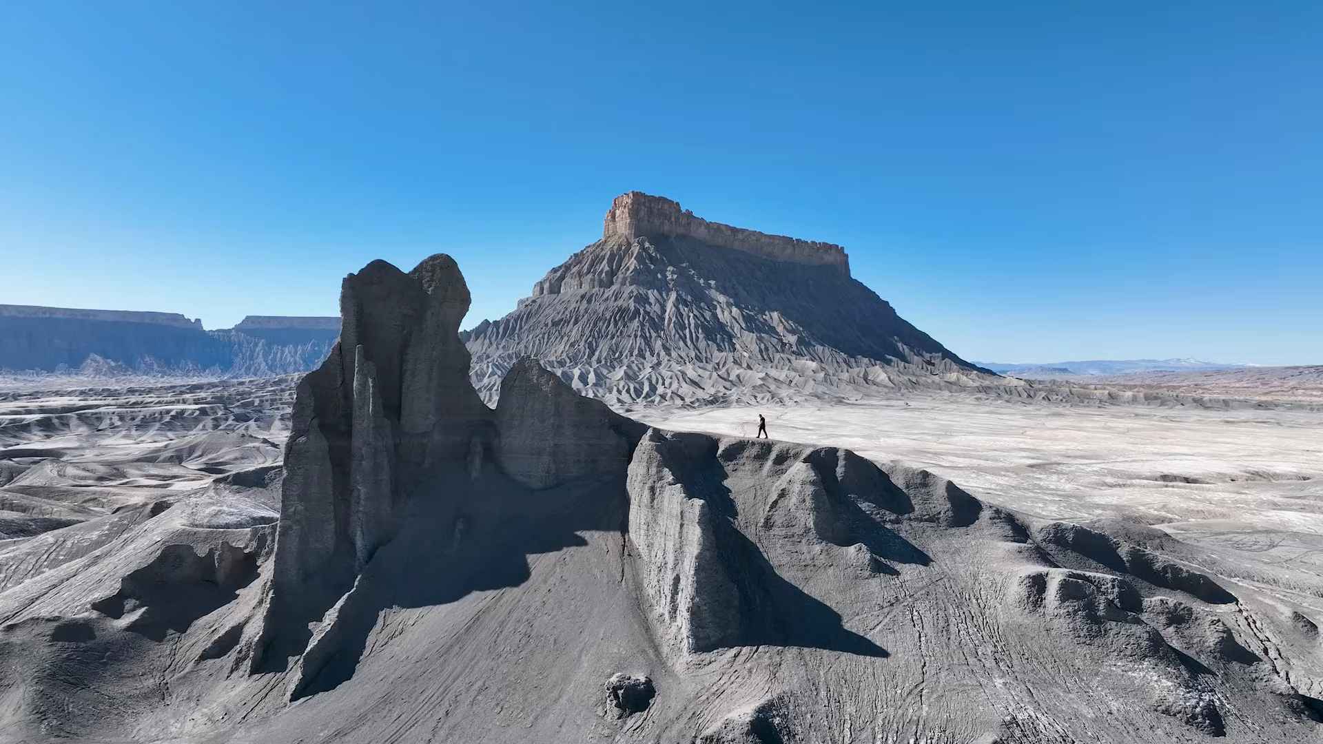Gray eroded hills with Factory Butte towering in the background under a clear blue sky