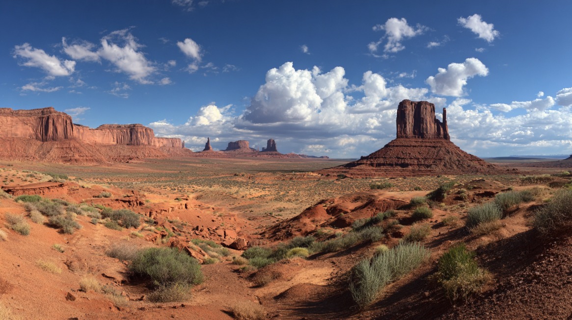 Panoramic desert scene with red sandstone buttes and mesas under a bright blue sky with scattered clouds in Monument Valley
