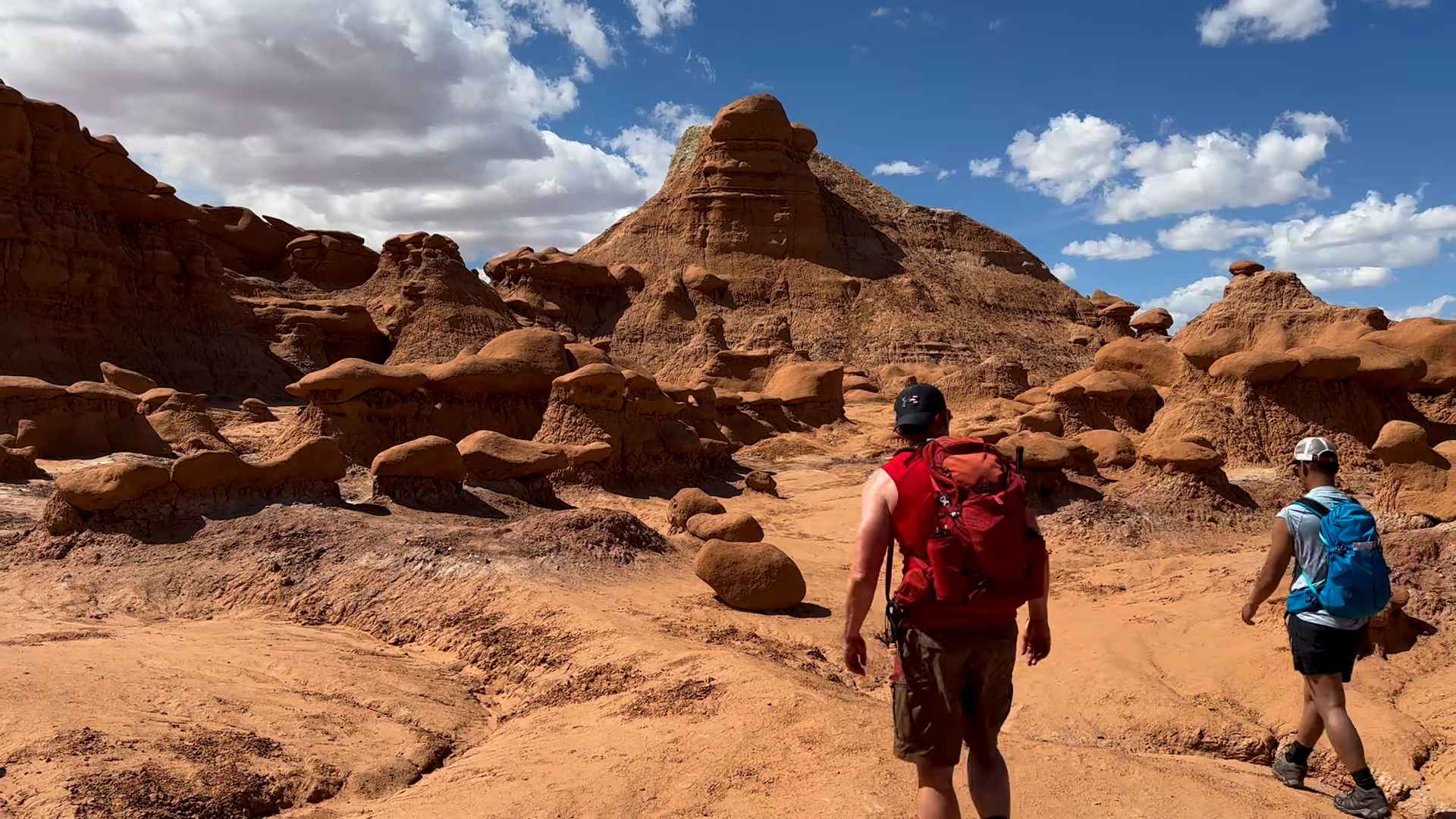 Two hikers with backpacks walking among mushroom shaped hoodoo rock formations in a desert landscape