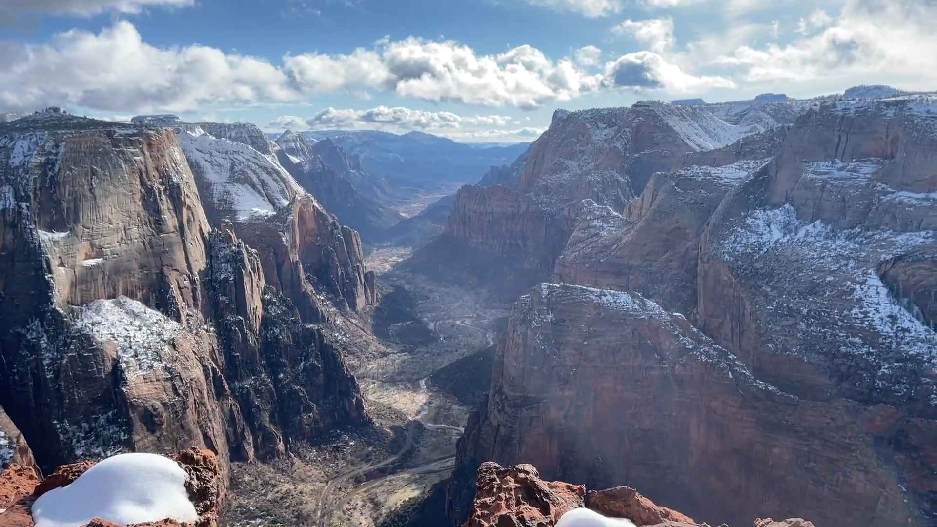 Wide canyon view with towering cliffs, a winding valley below, and patches of snow under a cloudy sky