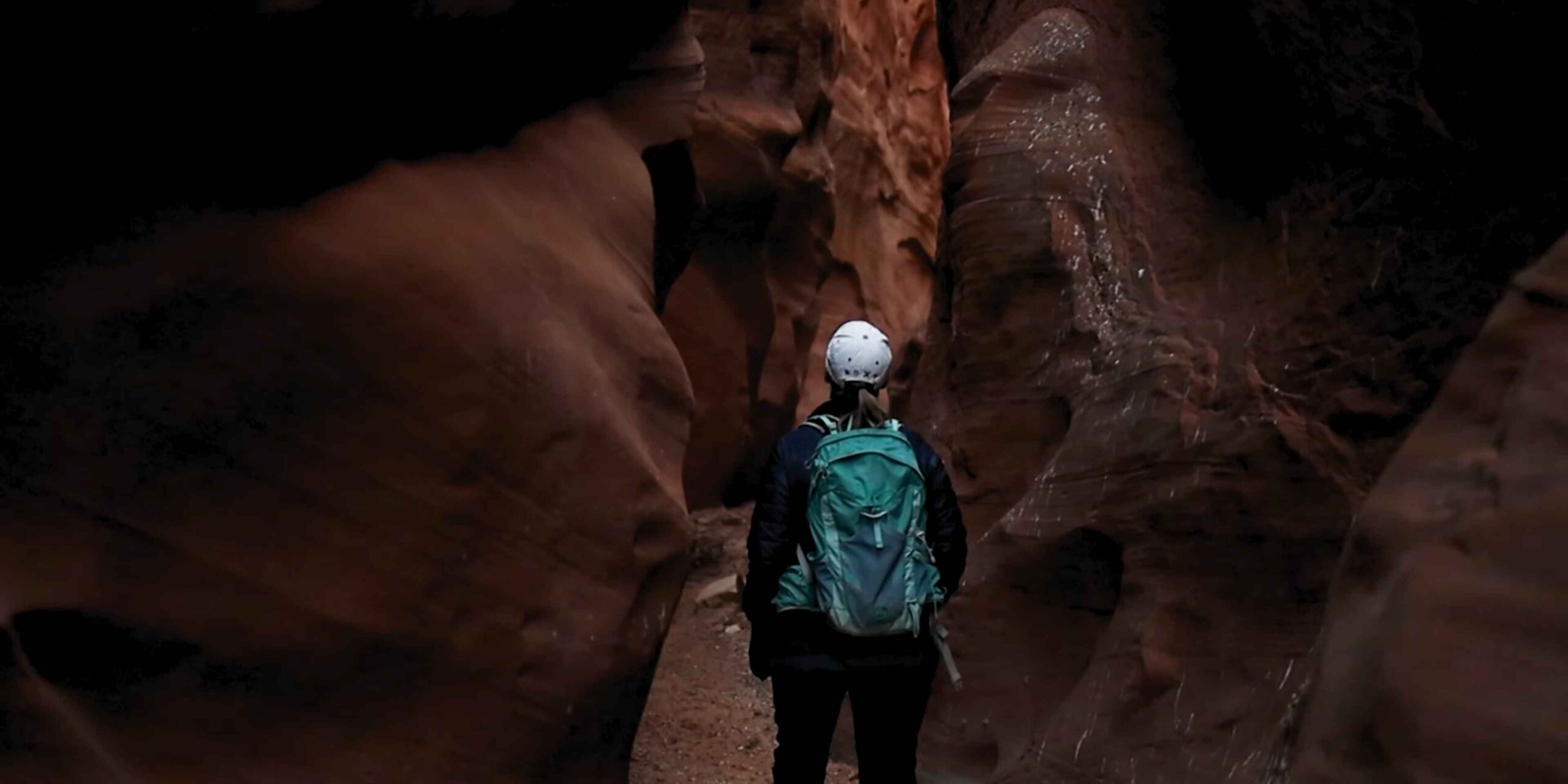 Hiker with a backpack walking through a narrow red rock slot canyon