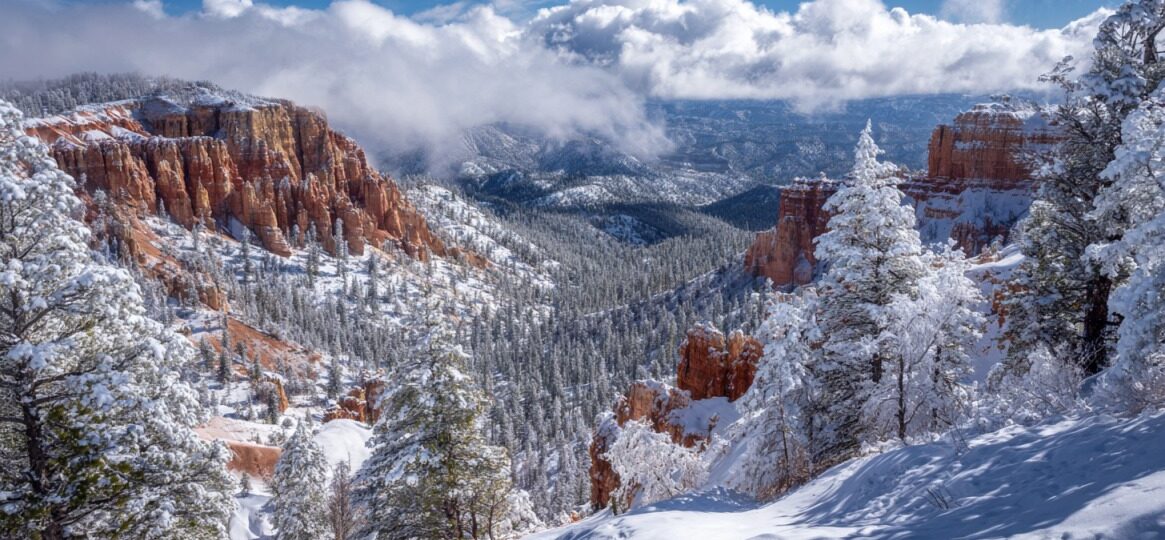 Snow-covered Bryce Canyon with orange hoodoos, frosted pine trees, and dramatic clouds filling the canyon under a bright winter sky.