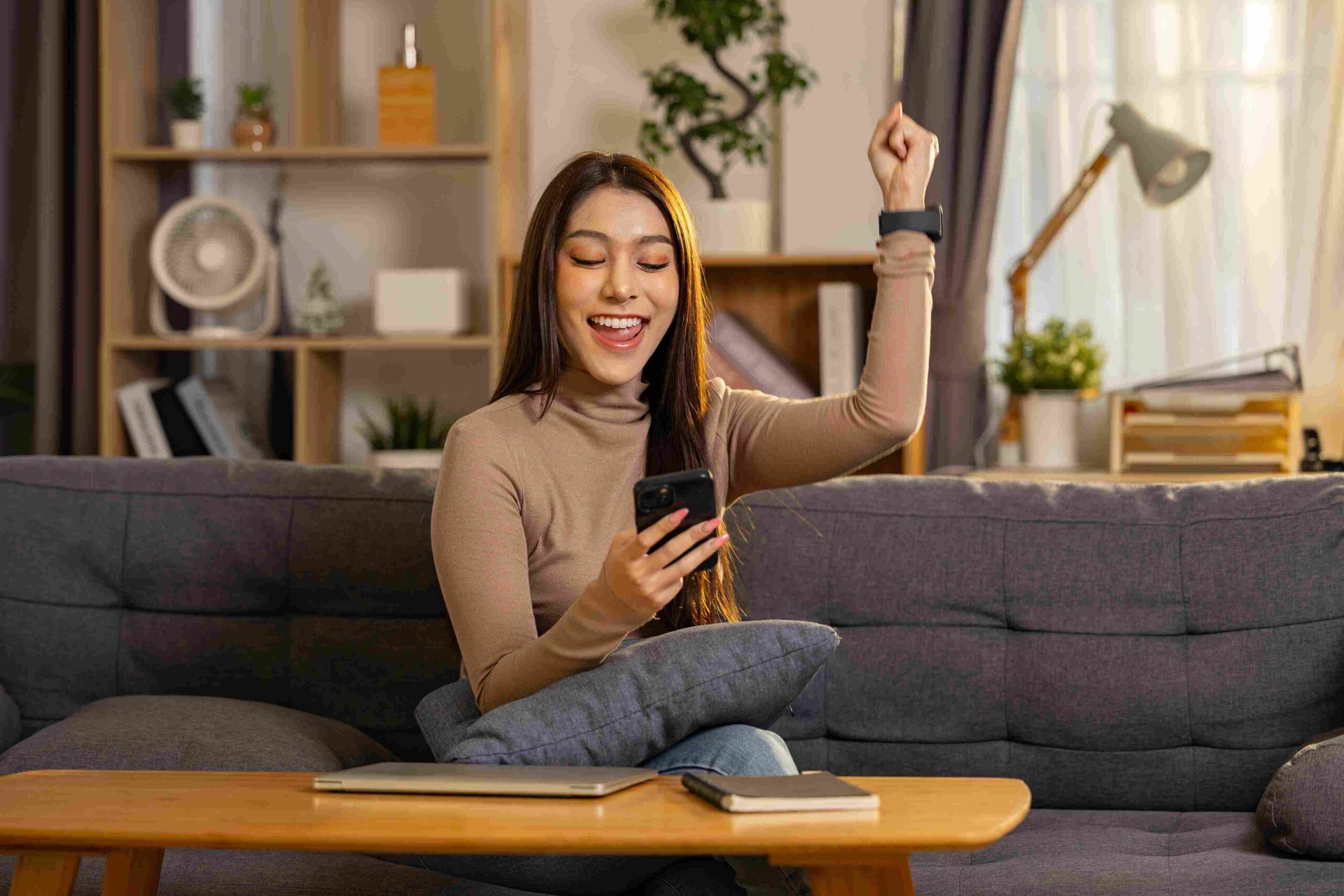 Woman sitting on a couch smiling and raising her fist while looking at her phone