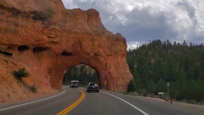 Cars drive through a rock tunnel along the Utah Mighty 5 Route with cliffs and forest in view