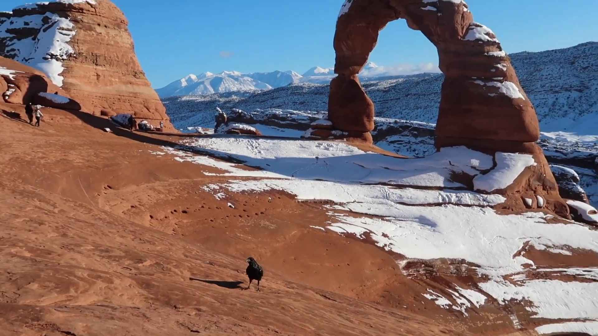 Natural rock arch in a red desert landscape with patches of snow and distant mountains