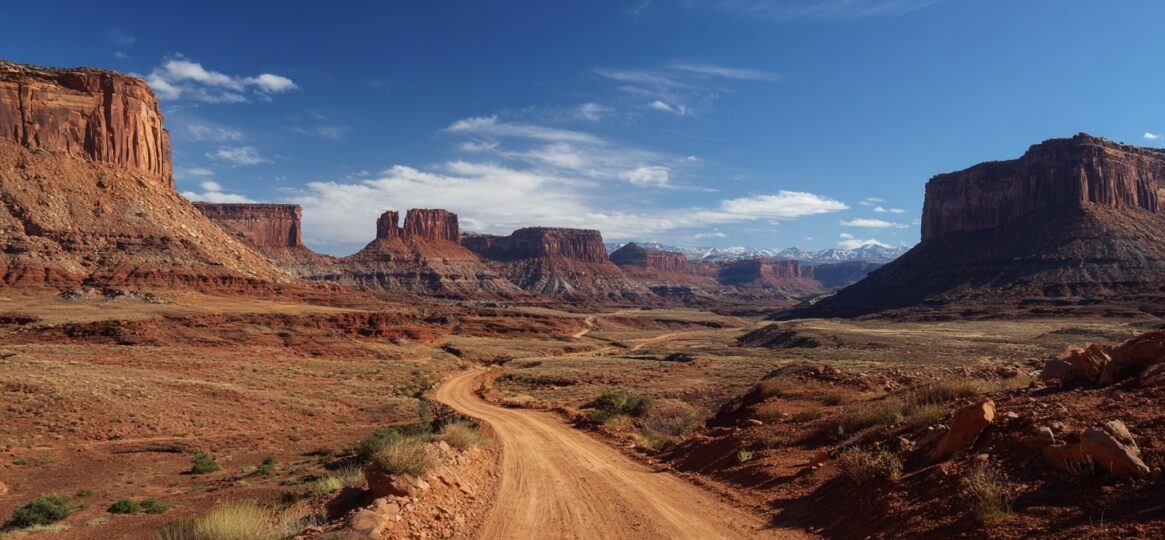 A winding dirt road stretching through a wide desert valley surrounded by massive red rock formations under a clear blue sky