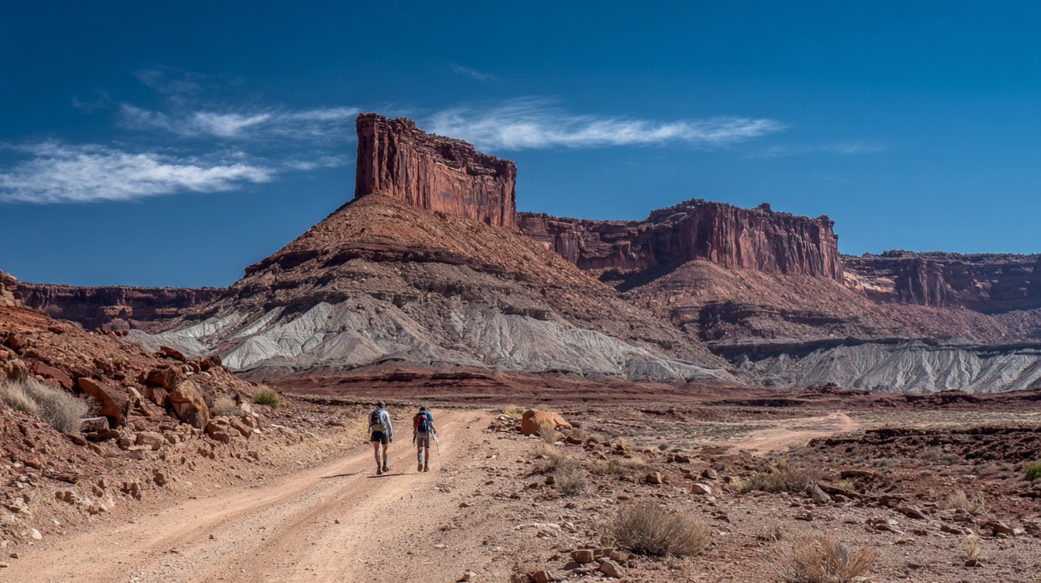 Two hikers walking along a dirt road surrounded by towering red rock formations and open desert terrain