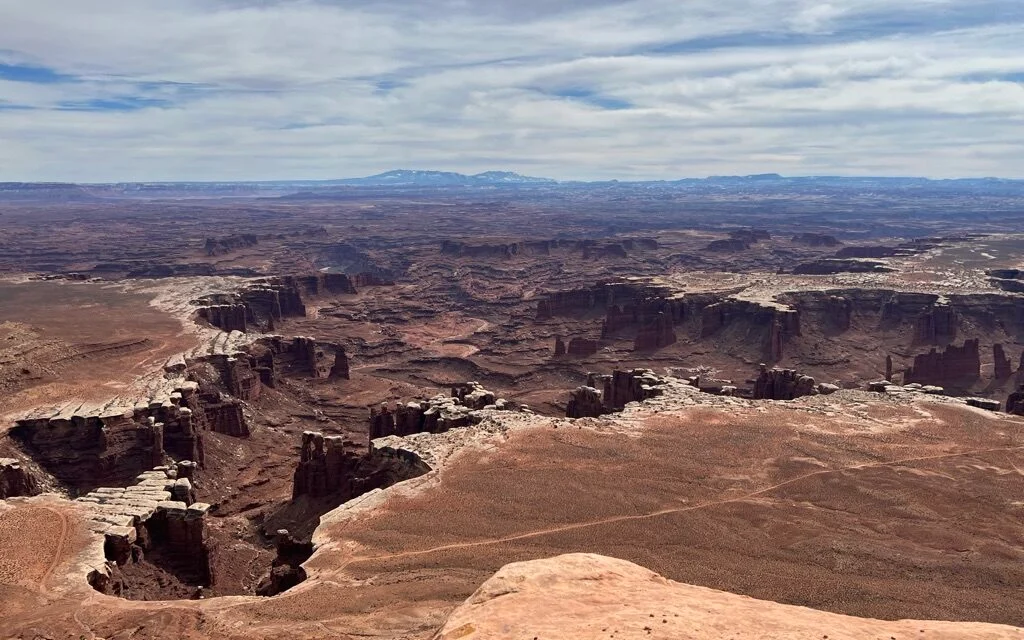 Aerial view of a dirt road cutting through a rocky desert with cliffs and distant mountains, with several vehicles traveling along it