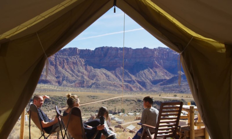 View from inside a tent looking out at four people relaxing in camp chairs, with vast, rugged mountains under a clear blue sky in the background