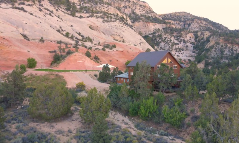 A cozy wooden cabin is nestled among green trees at the base of a rocky mountain with red and beige hues
