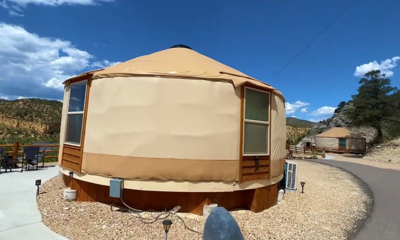 A beige yurt with large windows sits on a gravel path under a vibrant blue sky